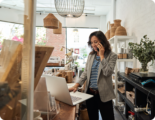 Home decor woman shop owner looking at laptop while on cell phone